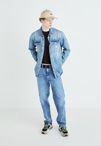 Gai zila džinsi jaka un atbilstoši džinsi, melns T-krekls zem tās, melna josta, beige cepure un daudzkrāsaini sporta apavi ar zaļiem akcentiem.