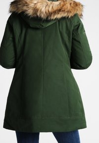 Mørkegrøn parka-jakke med et aftageligt faux fur hættefor. Den har en skræddersyet pasform og syninger, og længden når midt på låret.