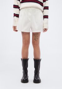 Λευκό σορτς από ελαφρύ ύφασμα με πιέτες στο μπροστινό μέρος, σε συνδυασμό με μαύρες δερμάτινες μπότες μέχρι το γόνατο. Πλεκτό πουλόβερ με υφή και ρίγες.