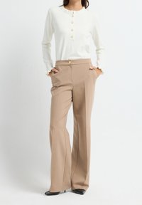Femme portant un haut blanc à manches longues avec des boutons et un pantalon large beige avec des poches, debout les mains dans les poches et chaussée de chaussures noires.