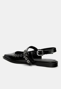 Svart spetsig slingback-sko med högblank läderfinish, prydd med cirkulära utskärningar och silverdetaljer på remmen vid ankeln.