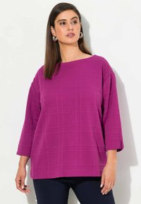 Fuchsia Oberteil mit einem strukturierten Karomuster, weitem Ausschnitt und dreiviertellangen Ärmeln, kombiniert mit dunkelblauen Hosen.