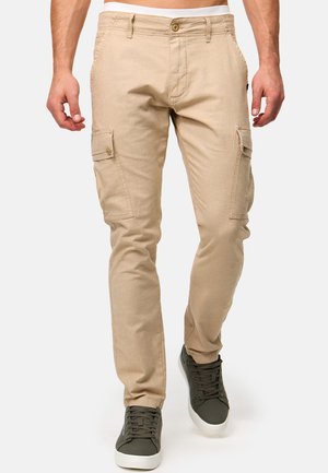 Pantaloni cargo beige con tasche laterali, design aderente e texture morbida. Indossati con sneaker verde scuro, caratterizzate da lacci frontali e suola in gomma.