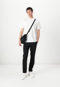 Valkoinen puuvillainen T-paita, jossa on vihreä logo, yhdistettynä mustiin housuihin ja valkoisiin lenkkareihin. Musta olkalaukku korostaa asua.