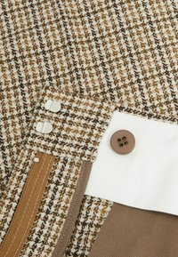 Tweedtyg med ett hundtandmönster i brunt, creamfärgat och beige. Har metalldetaljer och en brun knapp på en vit panel.