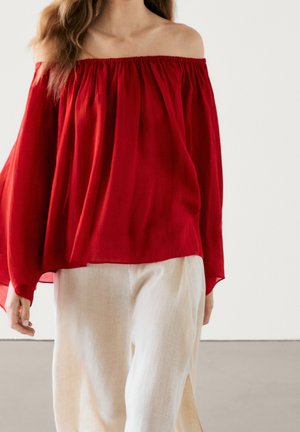 Woman wearing an off-shoulder loose red blouse paired with wide-leg light beige pants, standing against a plain background.