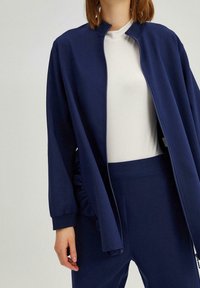 Marineblauwe zip-up jack met een hoge kraag, voorzien van gerimpelde accenten aan de zijkant, gedragen over een witte top en bijpassende broek.