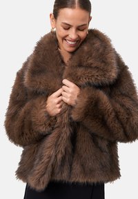 Veste en fausse fourrure marron avec un large col, texture douce et forme carrée, portée de manière lâche avec les mains tenant l'avant.
