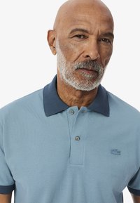 Ljusblå polo-shirt i texturerat tyg, marinblå krage och två-knappslå; har en broderad logotyp på vänster bröst.