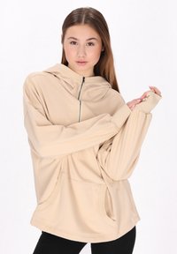 Beige színű kapucnis pulóver, puha anyagból készült, cipzáros záródással, elülső zsebekkel és hosszú ujjakkal, amelyeknél hüvelykujjnak nyílások találhatók.