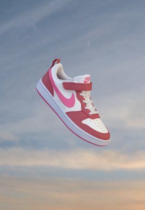 Basket Nike blanche et rose avec lacets et bande velcro flottant devant un ciel nuageux.