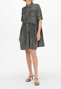 Harmaa denimmekko, jossa on lyhyet hihat, napitettava etuosa, kaksi rintataskua, kerätty vyötärö ja pehmeä tekstuuri; yhdistetty mustiin avoimiin kärkiin kengissä.