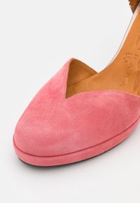 Chaussure en suede rose avec un design en V et un bout arrondi lisse. La doublure intérieure est en cuir marron, et la semelle est plate.