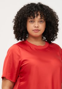 A woman with curly hair wears a red crewneck shirt, looking directly at the camera with a neutral expression against a light background.
