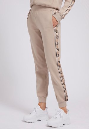 Pantalon jogger beige avec bande de marque sur les côtés, poignets et taille élastiques, porté avec des baskets blanches épaisses sur une personne debout.