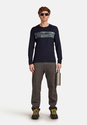 Langdurig mouwloze marineblauwe shirt met een gepatternd borstpaneel, grijze broek en zwarte schoenen met gele veters; houdt een zilveren waterfles vast.
