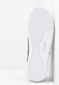 Semelle de chaussure en caoutchouc blanc avec des rainures horizontales et le nom de la marque "GUESS" embossé au centre, reposant contre un coin blanc.