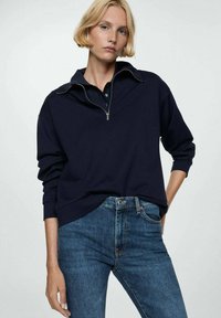 Pulover bleu marin cu guler cu fermoar pe jumătate și mâneci lungi, asortat cu blugi din denim albastru, având o croială clasică și detalii subtile de cusătură.