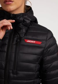 Giacca nera imbottita con chiusura a zip, cappuccio e una patch rossa con il logo "JACK1T", caratterizzata da una texture trapuntata e tessuto liscio.