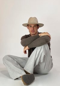 Beige Hut mit breiter Krempe, dunkelbrauner Pullover, hellgraue, locker zulaufende Hose, sitzende Pose auf einem weißen Hintergrund, texturiertes Schuhprofil sichtbar.
