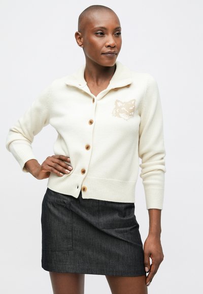 Fehér kötött kardigán sálgallérral, gombos záródással, a mellrészen barna róka mintával, sötét farmer szoknyával párosítva.