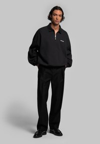Pull noir avec un col à zip quart, présentant une texture de tissu unie et un petit logo sur la poitrine, associé à un pantalon et des chaussures noirs.