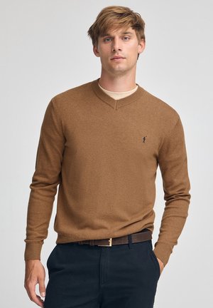 Bruine v-hals trui gemaakt van zacht materiaal, met een subtiel logo op de borst, gecombineerd met donkere broek en een bruine riem.