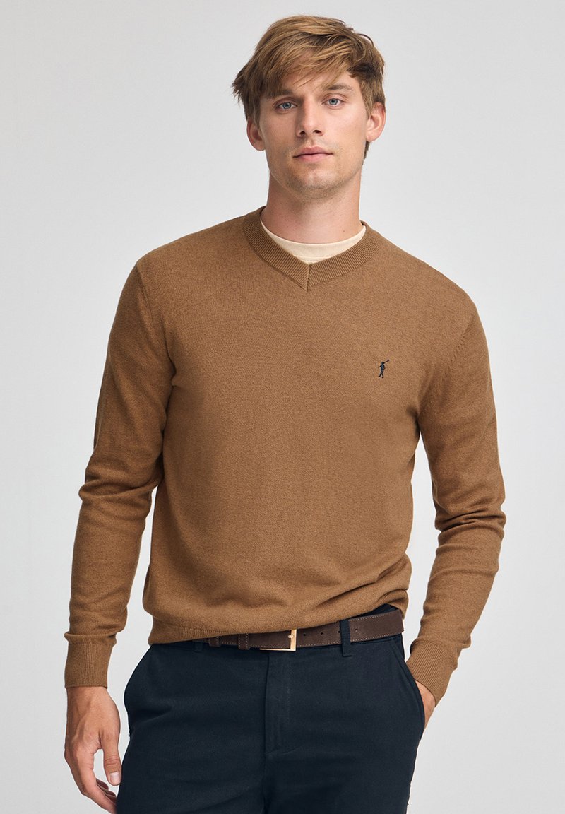 Brun v-ringad tröja i mjukt material med en diskret logga på bröstet, kombinerad med mörka byxor och ett brunt bälte.