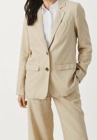 Mujer vestida con un traje de lino beige, con un blazer abotonado y pantalones de pierna ancha a juego, camisa blanca con cuello debajo, una mano en el bolsillo.