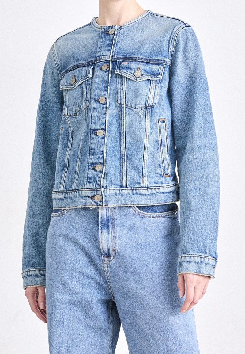 Chaqueta de mezclilla azul claro con cuello redondo, cierre de botones y dos bolsillos en el pecho. Presenta un ajuste relajado y una textura desgastada.