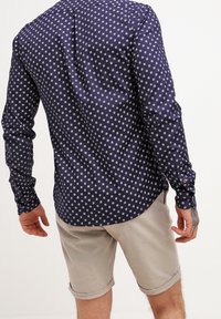 Camisa de manga larga en color marino con lunares blancos, con un collar abotonado y un dobladillo curvado. Combinada con pantalones cortos de color claro.