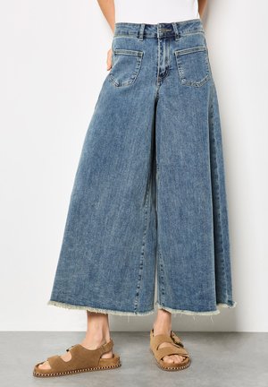 Pantalon denim à jambes larges en bleu clair avec un ourlet effiloché. Dispose de deux poches avant et d'une fermeture à bouton classique. Associé à des sandales marron.