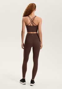 Barna sportos rövid felső, keresztpántokkal a háton, magas derekú barna leggingsszel párosítva. A modell fekete sportcipőt visel.