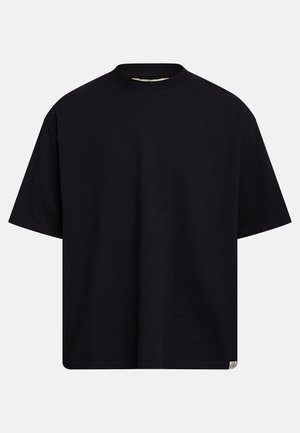 Fekete, túlméretezett rövid ujjú, kerek nyakú póló, alján egy kis törtfehér téglalap alakú címkével, világosszürke háttéren.