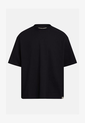 Fekete, túlméretezett rövid ujjú, kerek nyakú póló, alján egy kis törtfehér téglalap alakú címkével, világosszürke háttéren.