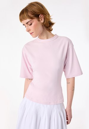Vrouw met kort lichtbruin haar, gekleed in een pastelroze top met korte mouwen en een witte geplooide rok, kijkt naar beneden naar haar linkerzijde.