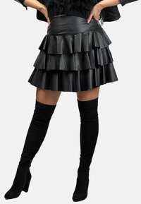 Czarna spódnica z sztucznej skóry z warstwowymi falbanami, dopasowanym paskiem i długością sięgającą do połowy uda. W połączeniu z wysokimi czarnymi skarpetkami i szpilkami.