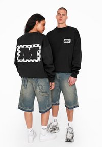 Schwarze Sweatshirts mit großen Logo-Patches; eines vorne, eines hinten. Getragen mit zerrissenen Jeansshorts, verziert mit Nähdetails.