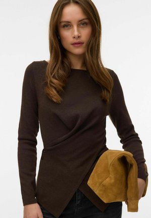 Joven mujer con cabello largo y castaño lleva una camiseta de manga larga marrón oscuro y sostiene una chaqueta de ante color beige doblada sobre un fondo blanco.