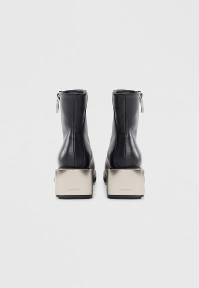 Crne kožne gležnjače s glatkom površinom, s niskim zatvaračem straga i metalnim blok potpeticama s ugraviranim brendom.