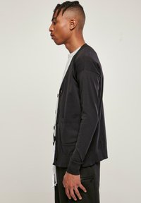 Fekete kardigán hosszú ujjakkal, elülső zsebekkel és gombos záródással. Puha anyagból készült, laza fazonban és bordázott részletekkel.