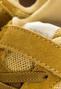 Sneaker in suede beige con una toma in rete e dettagli dorati, una tracolla testurizzata e lacci marroni. Cuciture strutturate e dettagliate sono ben visibili.