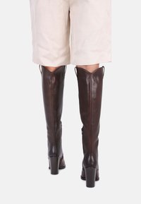 Bottes en cuir marron jusqu'à la cuisse avec une finition brillante. Elles présentent un bout rond, un talon bloc haut et des zips au dos. Extérieur texturé.