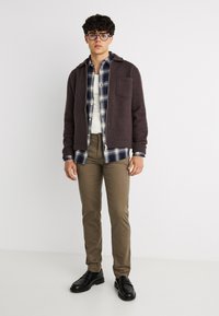 Νεαρός άνδρας με γυαλιά, καφέ μπουφάν με φερμουάρ, μπλε καρό πουκάμισο πάνω από λευκό μπλουζάκι, χακί παντελόνια και μαύρα παπούτσια, στέκεται μπροστά σε λευκό φόντο.