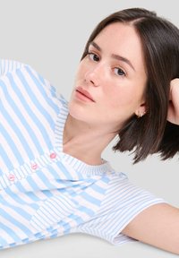 Top de manga corta a rayas azul y blanco con escote en V, que cuenta con botones rosas y un bolsillo decorativo. Tejido de algodón suave.