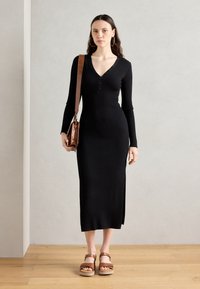 Black ribbed long-sleeve dress with a V-neck, button detail, and side slit. Paired with brown platform sandals and a textured brown handbag.