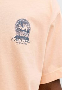 Tricou de culoare piersică deschisă cu un emblemă bleumarin reprezentând un cal în poziție de trăsnet și textul „The IVY Manor originals ©1990” pe piept.