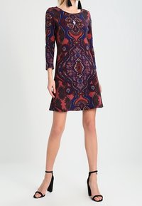 Květinové šaty s dlouhým rukávem v červené, modré a černé barvě, s kulatým výstřihem a rozšířeným lemem, doplněné černými sandály na podpatku.