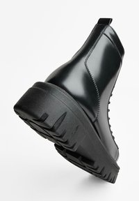 Černé kotníkové boty z lesklé kůže, s tlustou černou gumovou podrážkou s texturovaným vzorem běhounu a poutkem na zadní straně.