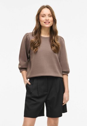 Femme aux longs cheveux bruns ondulés portant un haut marron à manches bouffantes et un short noir plissé, debout avec une main dans la poche sur fond blanc.